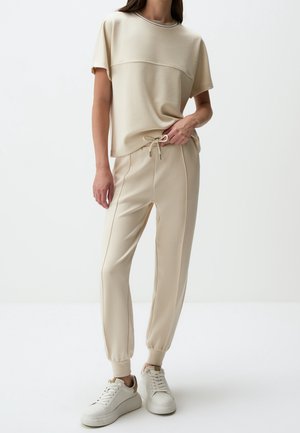 Kobieta ubrana w beżową bluzkę z krótkim rękawem oraz pasujące joggery ze ściągaczem i białe trampki, stojąca na jednolitym białym tle.