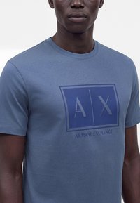 Άνδρας που φορά μπλε μπλουζάκι Armani Exchange με μεγάλο λογότυπο AX και το όνομα της μάρκας στο στήθος, σε απλό λευκό φόντο.