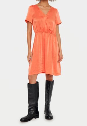 Femme portant une robe orange courte à manches courtes et longueur genoux avec un col en V et des bottes en cuir noires montant jusqu’aux genoux sur un fond uni.
