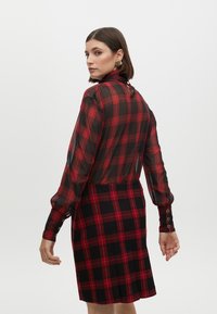 Punainen ja musta ruudullinen mekko, jossa on läpinäkyvä yläosa, pitkät hihat ja napituskoristeet hihoissa, varustettuna tiukalla hameella.