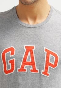 Camiseta de algodón gris con letras "GAP" bordadas en rojo, escote redondo y mangas cortas, con una textura suave.