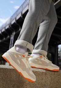 Ljusgrå sneakers med orange accenter, i mocka och nätmaterial, rund tå, vadderad krage och vita snören. Bärs med uppvikta grå byxor.