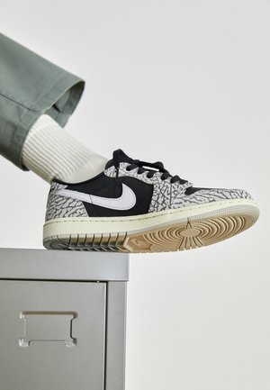 Stopa w czarno-szarym bucie marki Nike z białym logo, oparta na krawędzi szarej metalowej szafki, na jednolitym tle.