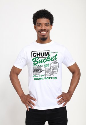 Uomo sorridente con capelli ricci indossa una maglietta bianca con la scritta "Chum Bucket, Bikini Bottom", che elenca gli elementi del menu e presenta una grafica di un secchio da cartone animato sul davanti.
