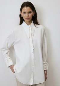 Witte lange mouwen shirt met een hoge kraag, sluiting met knopen aan de voorkant en koorden bij de manchetten. Gemaakt van zachte, gladde stof met een relaxed fit.