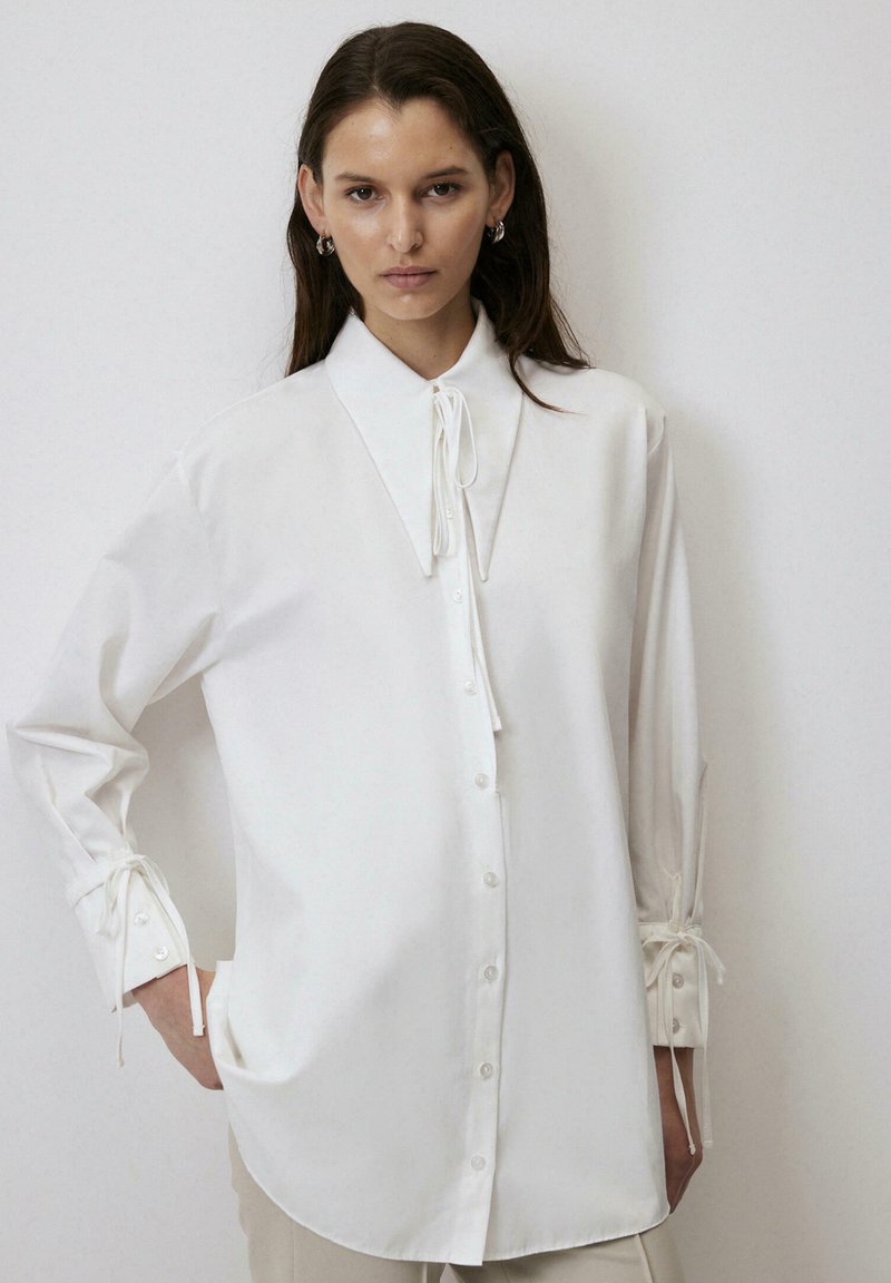 Witte lange mouwen shirt met een hoge kraag, sluiting met knopen aan de voorkant en koorden bij de manchetten. Gemaakt van zachte, gladde stof met een relaxed fit.