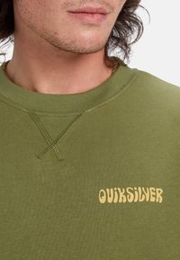 Olive zelená mikina vyrobená z mäkkej tkaniny, s okrúhlym výstrihom, viditeľným šitím a žltým logom "Quiksilver" na prednej strane.
