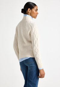 Cremefarvet strikket sweater med teksturerede mønstre, med en høj hals og ribbede manchetter, båret over en blåstribet skjorte med krave.