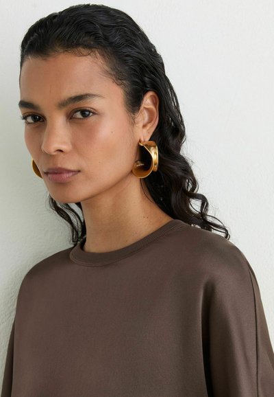 Woman with dark wavy hair wearing oversized gold hoop earrings and a brown crew neck top, looking slightly to the side against a white background.