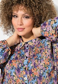 Košile s květinovým vzorem, která má límeček, zapínání na knoflíky a pletenou texturu. Barvy zahrnují fialovou, oranžovou, růžovou a modrou.