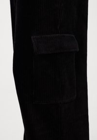 Zwarte corduroybroek met verticale ribbeling, voorzien van een zijzak met een klep. De stof heeft een zachte textuur en een matte afwerking.