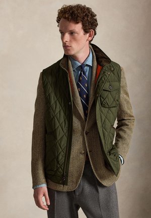 Man met krullend haar die een olijfgroene doorgestikte bodywarmer draagt over een beige blazer met visgraatpatroon, een blauw overhemd, een marineblauwe stropdas met patroon en grijze broek.
