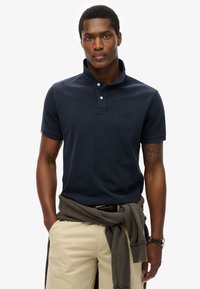 Mørkeblå polo shirt med krave, tre knapper og broderet logo. Lette strukturerede materialer med en tætsiddende pasform, parret med khaki shorts.