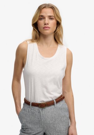 Femme blonde portant un haut blanc sans manches, un pantalon rayé taille haute et une ceinture tressée marron, debout avec une main dans la poche.