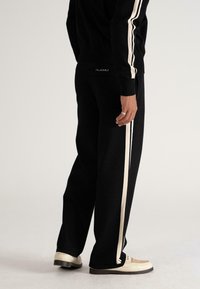Person wearing black pants and jacket with white side stripes, standing, beige shoes visible on a light background.
