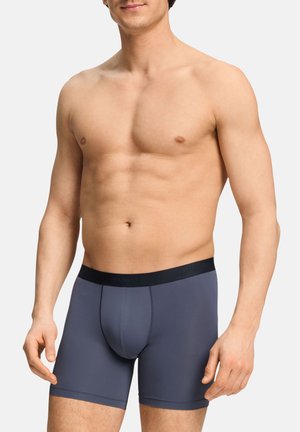 Muscular man wearing navy blue boxer briefs with black waistband, standing against a plain light background.