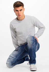 Sweatshirt gris à manches longues associé à un jean en denim bleu avec des motifs distressed et des détails de couture, porté avec des baskets blanches.