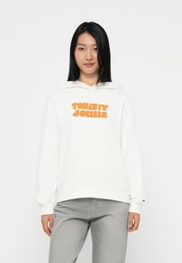 Felpa bianca con logo arancione "TOMMY JEANS" sul davanti, con cappuccio regolabile e polsini a coste. Abbinata a jeans grigi.