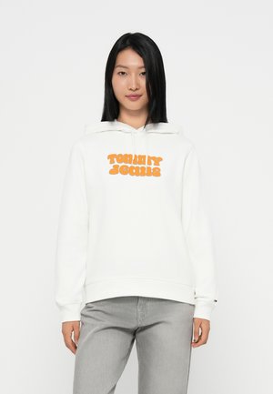 Felpa bianca con logo arancione "TOMMY JEANS" sul davanti, con cappuccio regolabile e polsini a coste. Abbinata a jeans grigi.