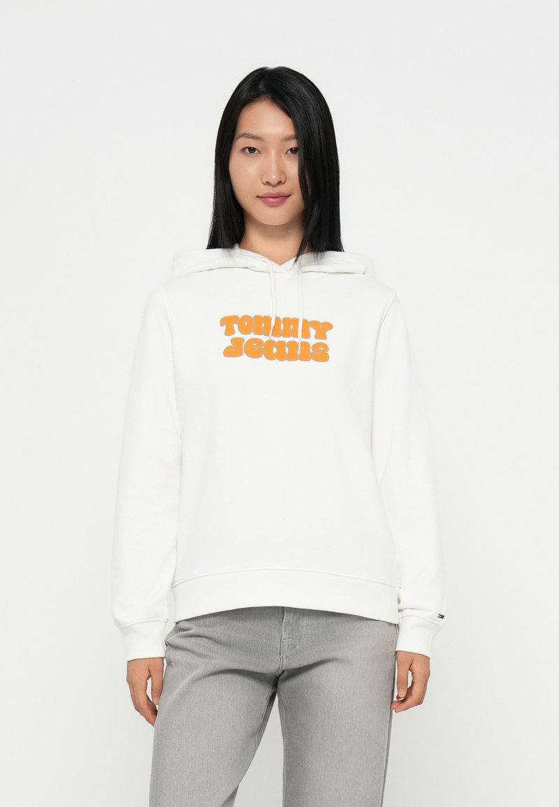 Felpa bianca con logo arancione "TOMMY JEANS" sul davanti, con cappuccio regolabile e polsini a coste. Abbinata a jeans grigi.