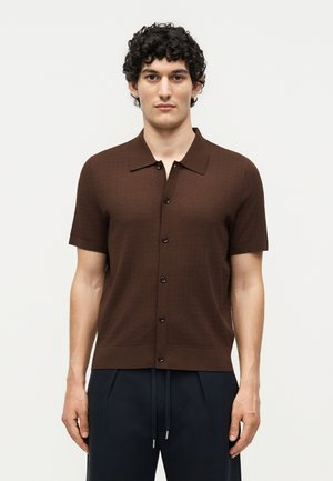 Νέος άνδρας με σγουρά σκουρόχρωμα μαλλιά, φορώντας ένα καφέ κοντομάνικο πουκάμισο με κουμπιά και μπλε σκούρο παντελόνι με κορδόνι, στέκεται μπροστά σε απλό φόντο.