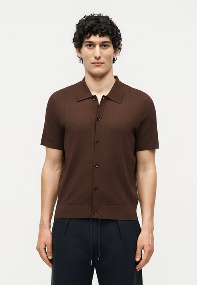 Young man with curly dark hair wearing a brown short-sleeve button-up shirt and navy drawstring pants, standing against a plain background.