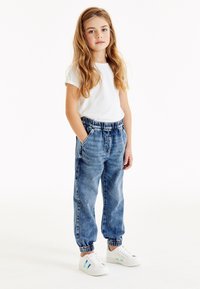 Joger denim dengan pinggang elastis, dua kantong samping, dan cuffs bergaris, dipasangkan dengan kaos putih lengan pendek dan sepatu kets putih.