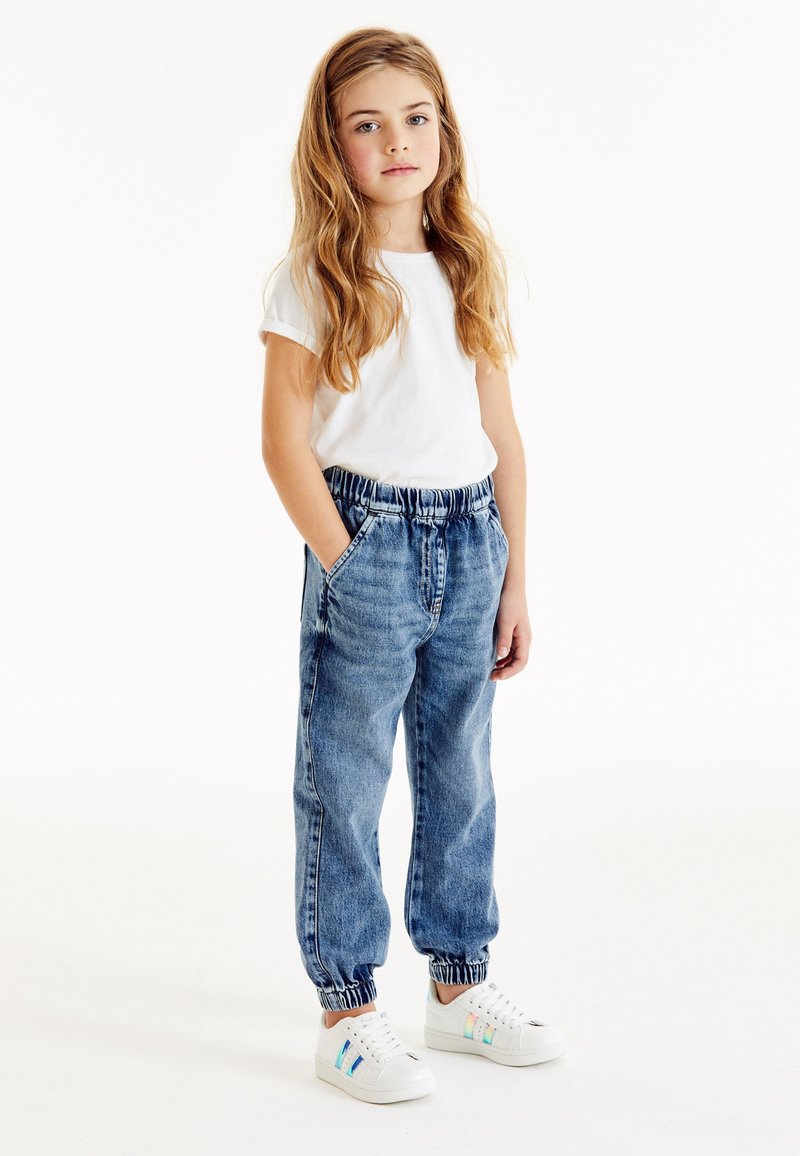 Joger denim dengan pinggang elastis, dua kantong samping, dan cuffs bergaris, dipasangkan dengan kaos putih lengan pendek dan sepatu kets putih.