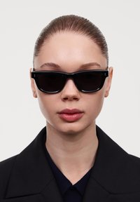 Čierne slnečné okuliare s hrubými štvorcovými rámami a tmavými sklami, nosené osobou s elegantným účesom, na neutrálnom pozadí.