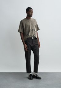 Olijfgroene oversized T-shirt met gedrukte tekst, gecombineerd met donkere aansluitende broek en zwarte leren schoenen. Soepele textuur, casual ontwerp.
