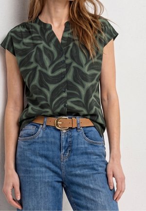 Vrouw draagt een mouwloze donker groene blouse met bladmotief en knopen, blauwe spijkerbroek en een beige riem met een gouden gesp.