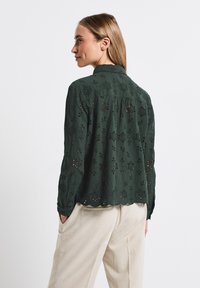 Donkergroen blouse met bloemborduursels en uitsparingen, met een kraag en een gegolfde zoom. Dragen over lichtbeige broeken.