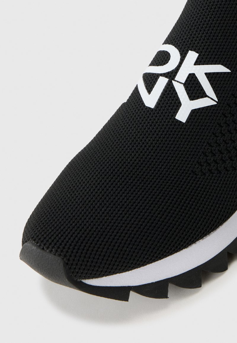 Baskets noires en tissu tricoté texturé, avec un logo blanc proéminent et une semelle contrastante noire et blanche aux bords dentelés.