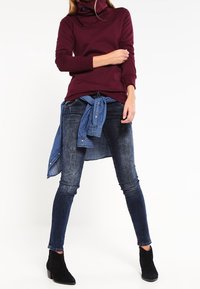 Sudadera burdeos con cuello de cowl, camisa de mezclilla azul amarrada a la cintura, jeans skinny de lavado oscuro y botas negras hasta el tobillo. Tela suave y diseño ajustado.