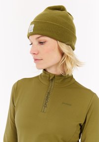Vrouw met kort blond haar, gekleed in een olijfgroen beanie en een bijpassend ritsjack, die naar beneden en opzij kijkt tegen een witte achtergrond.