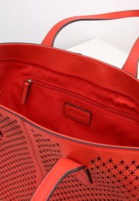 Sac fourre-tout rouge en matériau synthétique, avec un design perforé complexe, doublure intérieure lisse et une petite poche zippée.