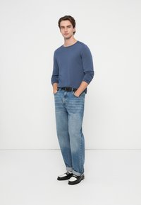 Niebieska koszula z długim rękawem, jasnoniebieskie dżinsy, czarny pasek i czarne mokasyny z białymi akcentami. Model stoi z rękami w kieszeniach.