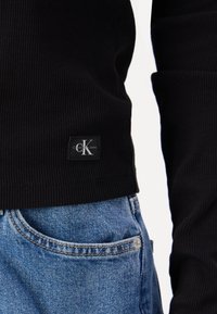 Μαύρος ριμπ πουλόβερ με ετικέτα λογότυπου στο στρίφωμα, φορεμένο πάνω από μπλε τζιν παντελόνι με ορατή τσέπη και λεπτομέρεια ραφής.