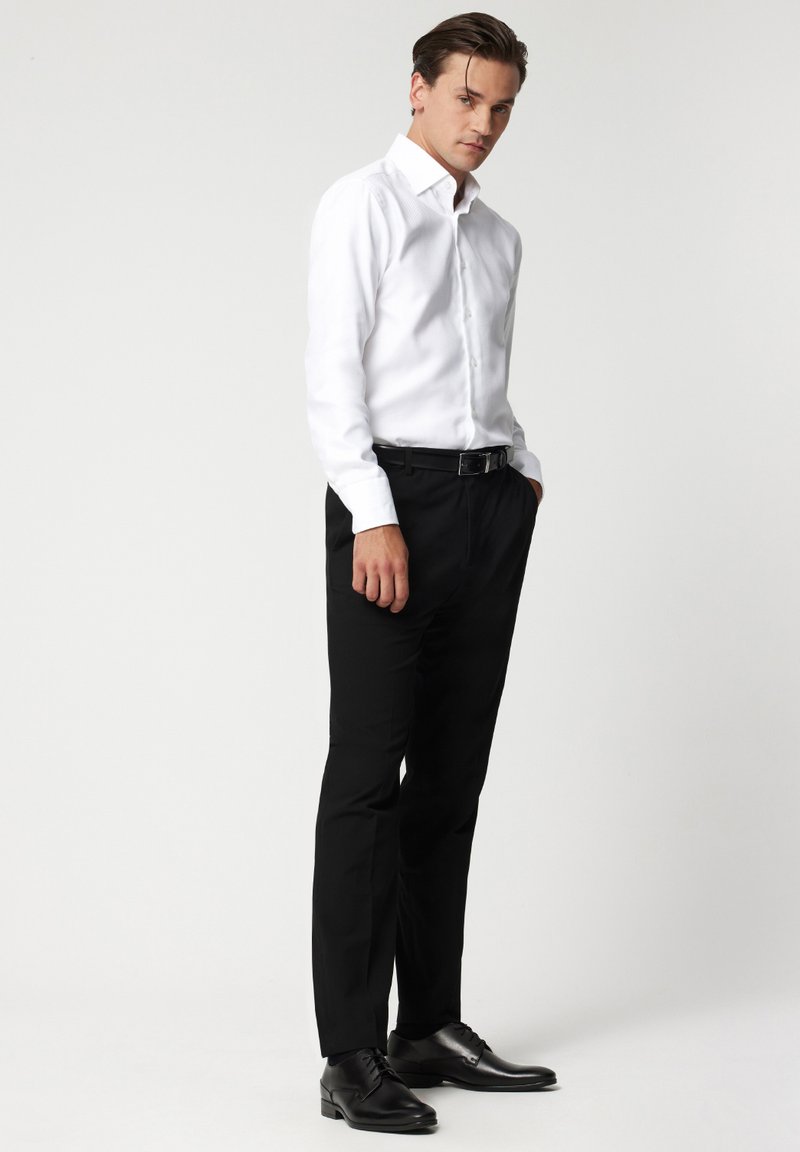 Biała koszula z długim rękawem i kołnierzykiem zapinanym na guziki, w zestawieniu z dopasowanymi czarnymi spodniami i czarnymi butami wizytowymi. Prosty, dobrze dopasowany krój.