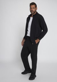 Giacca nera con zip e colletto rialzato, indossata sopra una t-shirt bianca, abbinata a pantaloni della tuta neri e sneaker nere. Design semplice e sportivo.
