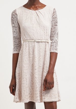 Femme portant une robe en dentelle beige clair avec des manches trois-quarts et une fine ceinture assortie à la taille.