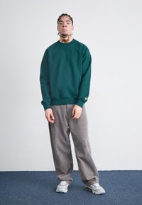 Grön sweatshirt med rund halsringning, ribbade ärmslut och en diskret logotyp på ärmen; kombinerad med ljusgrå manchesterbyxor och silvriga sneakers.