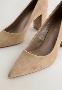 Escarpins beiges en daim à bout pointu avec talons carrés, inclinés pour montrer la doublure intérieure et les détails de couture sur un fond clair.