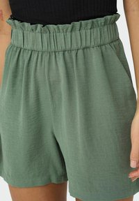 Calções verdes leves, feitos de tecido suave; apresenta uma cintura elástica com fru-fru e bolsos laterais; ajuste solto e fluido.