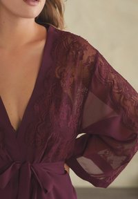 Robe bordeaux con delicate rifiniture in pizzo e maniche trasparenti, caratterizzata da un design avvolgente e una vita legata per una finitura morbida e testurizzata.