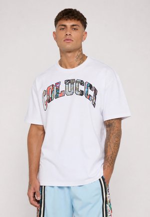 Mężczyzna z tatuażami ubrany w białą koszulkę z kolorowym napisem "COLUCCI" oraz jasnoniebieskie spodenki z wzorzystymi paskami po bokach, stojący na jednolitym tle.