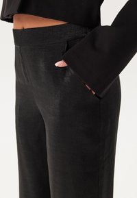 Zwarte hooggetailleerde broek van gladde stof, met zijzakken en een subtiele glans. Het ontwerp heeft wijde pijpen en tailorgedeelten.