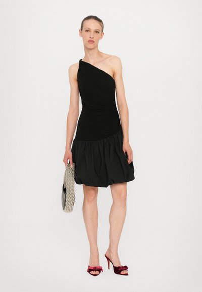 Robe noire à une épaule avec corsage ajusté et jupe évasée à volants. Le modèle tient une pochette argentée ornée de perles et porte des sandales à talons hauts en velours rouge.