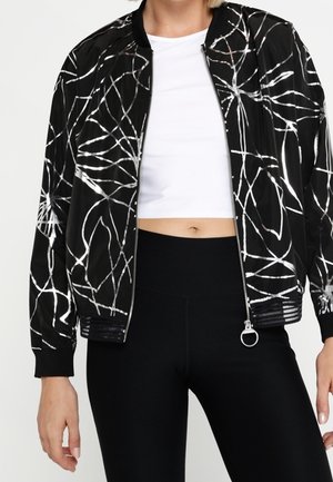 Frau trägt eine schwarze Bomberjacke mit weißen abstrakten Linien, ein weißes bauchfreies Top und schwarze Leggings mit hoher Taille.