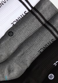 Três pares de meias caneladas nas cores branca, cinza e preta. Cada par apresenta listras contrastantes e detalhes de logotipo. Textura de tecido suave.